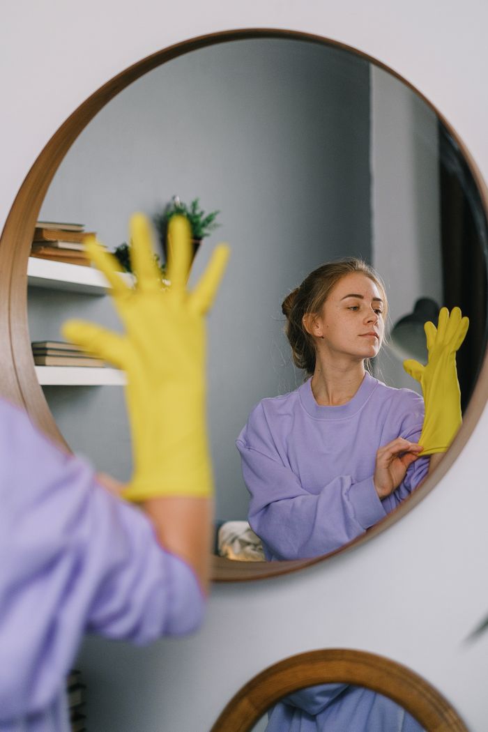 services-02 A woman wearing yellow gloves is reflected in a wall mirror indoors, exuding a serene expression.